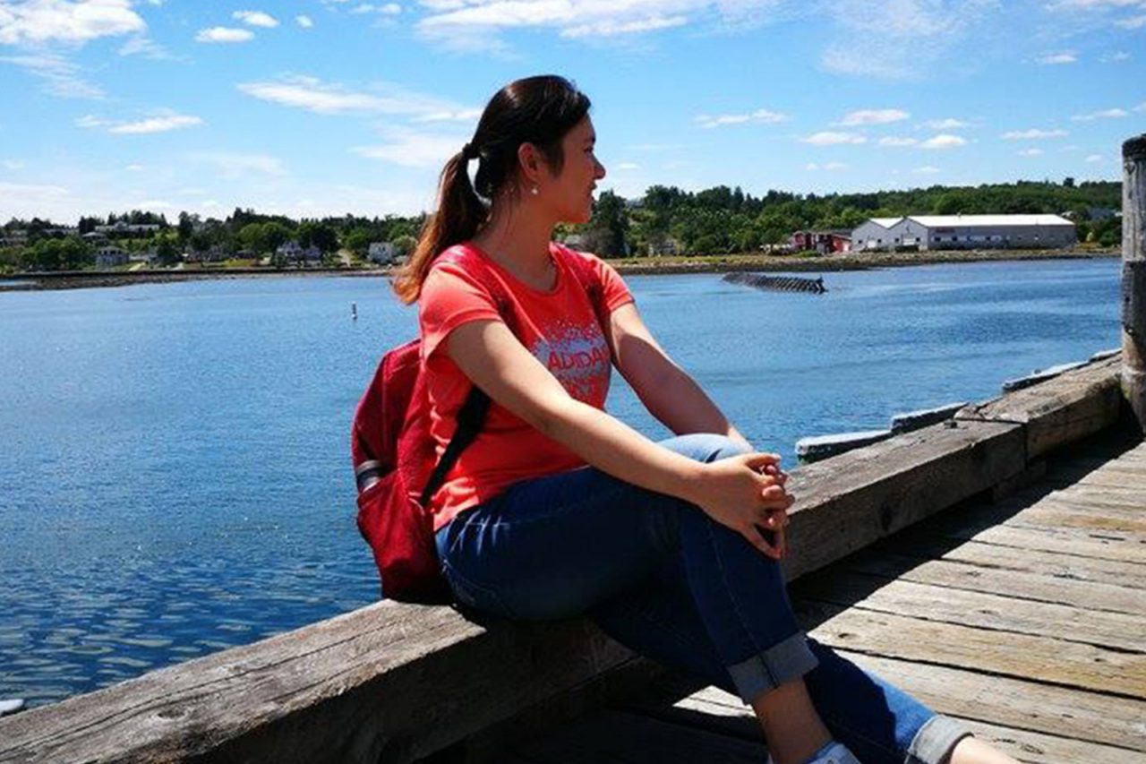 A female student posing on the wharf with the ocean and green fields in the background.
