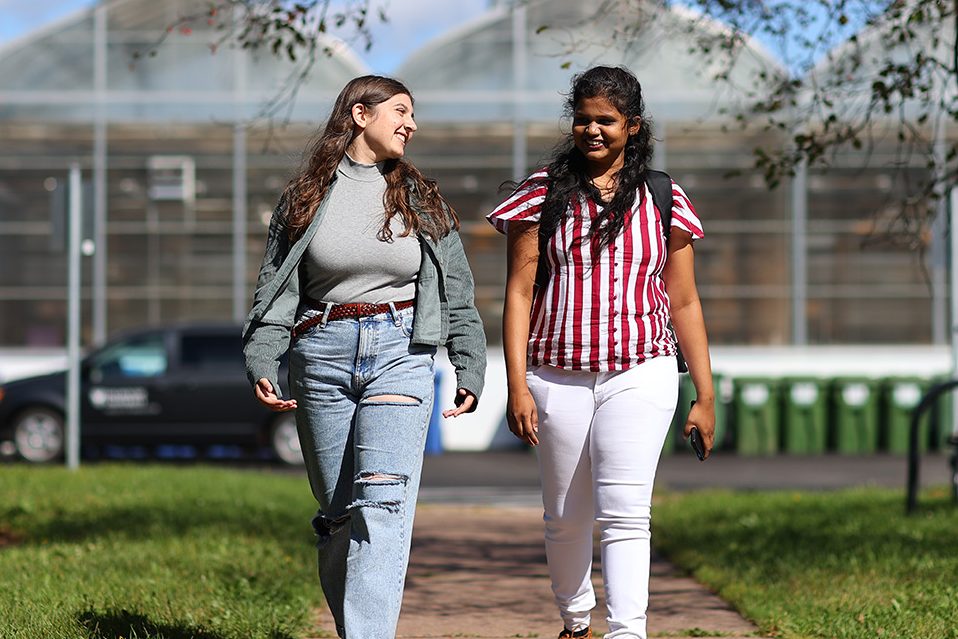 Two female students walking in front of a greenhouse.