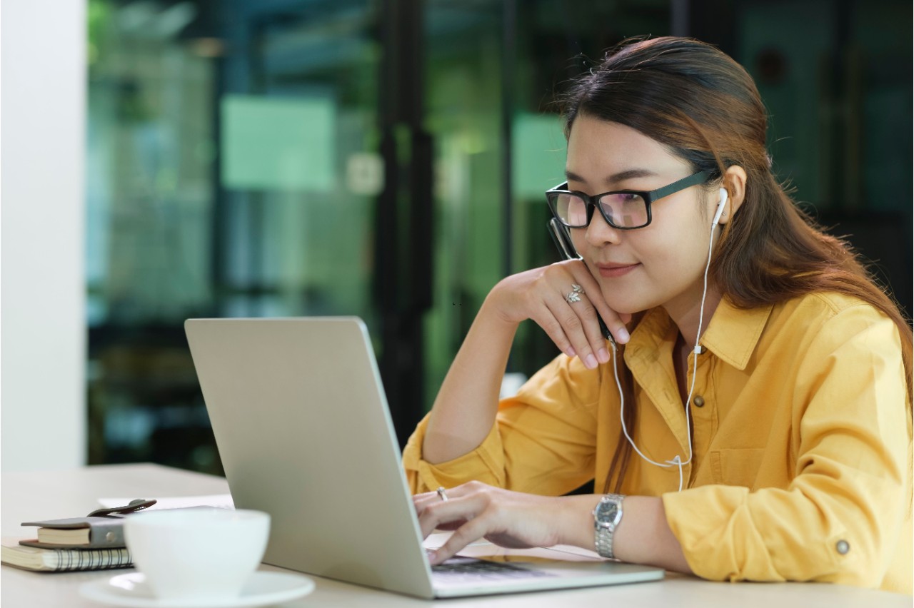 A young woman wearing a yellow shirt and glasses sitting at a desk with headphones in while working on her laptop.