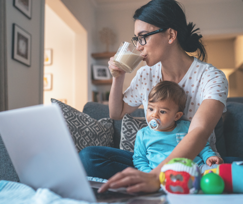 Woman wearing glasses working on a laptop drinking coffee with a baby on her lap.