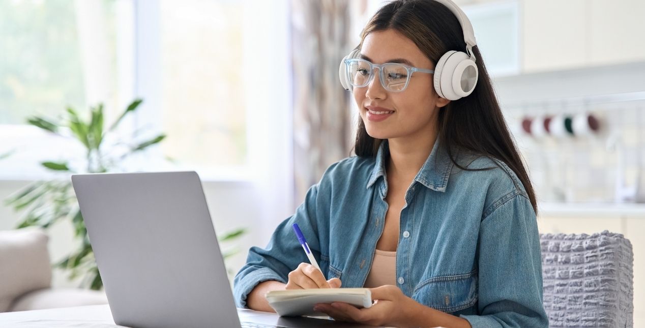 A student is wearing headphones while listening to a lecture on her computer and takes notes.