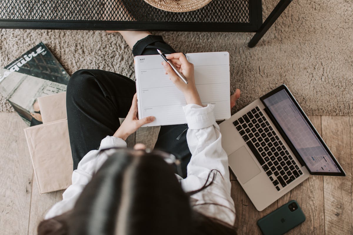 An overhead view of a student sitting on a carpet, surrounding by books, a laptop computer, and a mobile phone. They are writing in a notebook. Photo by Vlada Karpovich.