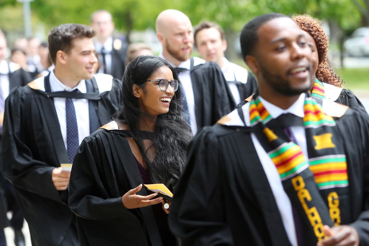 A group of graduating students wearing gowns walking in a group outdoors on a fine day on their way to an event.