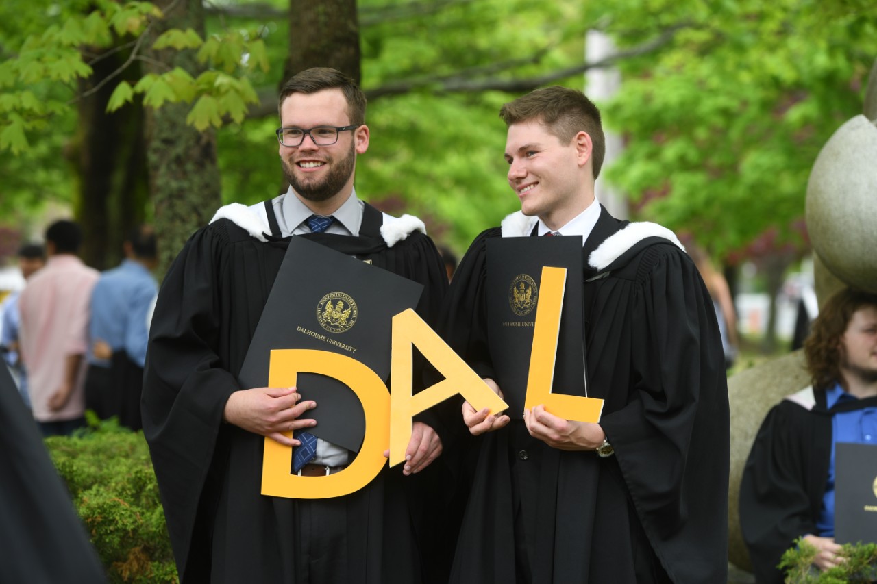 Two students wearing convocation robes hold large yellow letters spelling DAL.