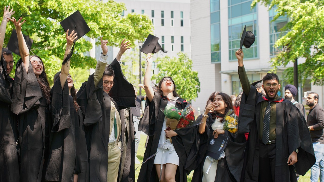A group of students wearing convocation gowns cheering and throwing their caps in the air.  