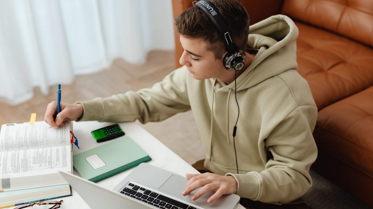 Student studying on lap top wearing headphones and consulting a text book.