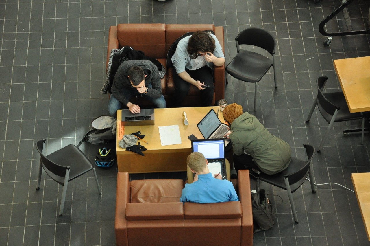Four students are shown from above sitting on chairs and sofas around a low table. Three are working on laptop computers and the fourth is looking at a mobile phone.