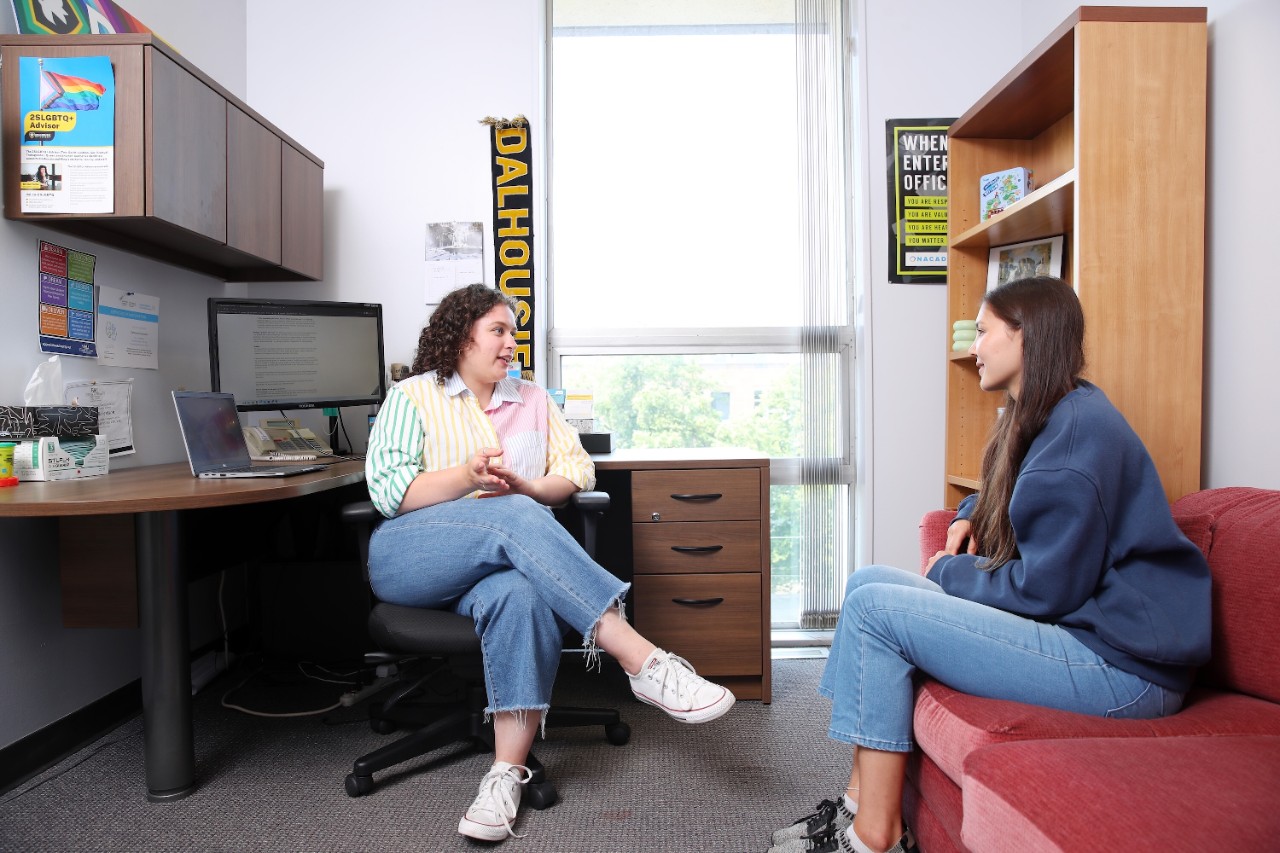 A Dalhousie advisor sits beside her desk while talking with a student who is sitting on a red sofa.