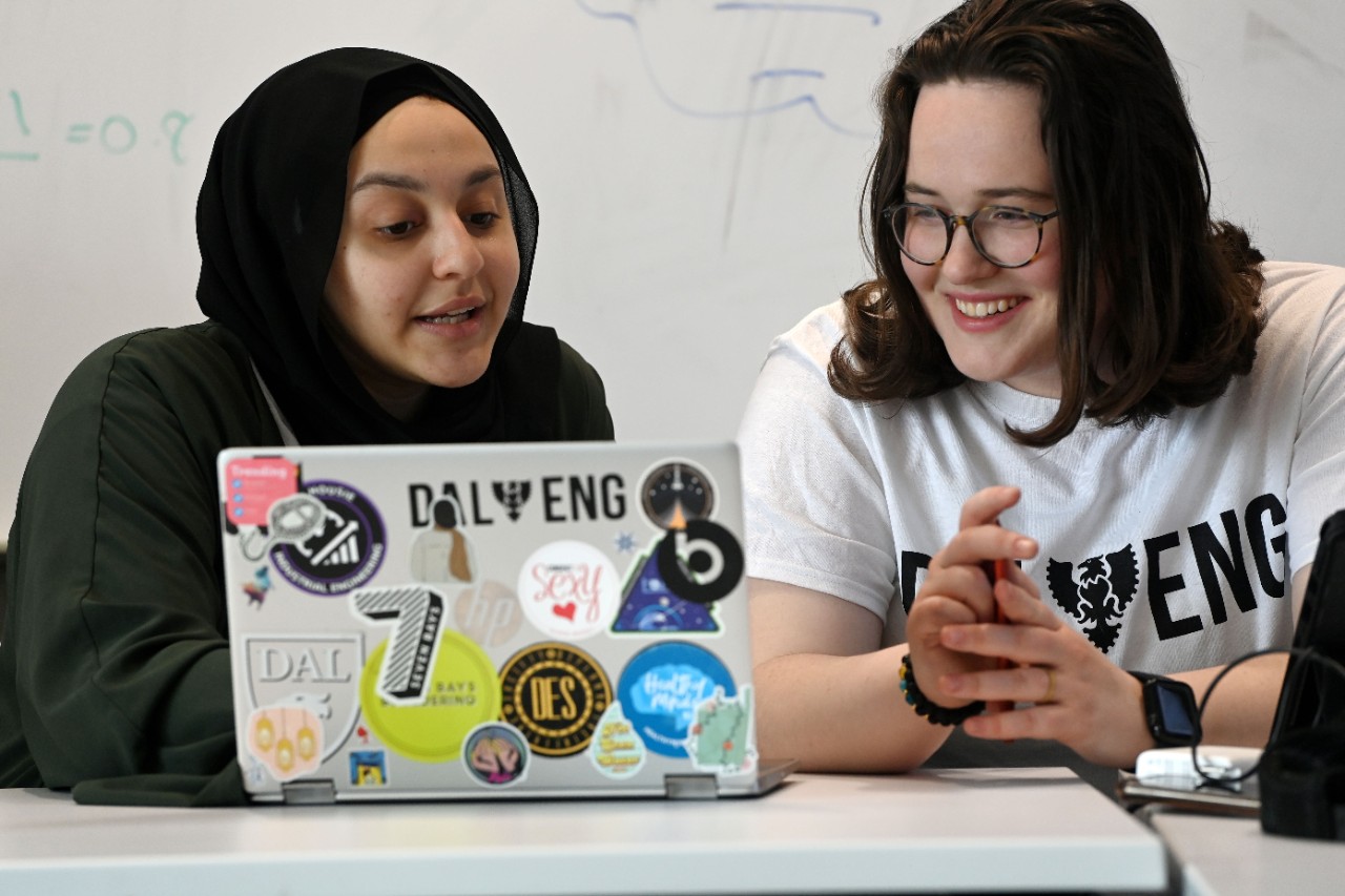2 Dal engineering students converse while looking at a laptop covered in stickers.