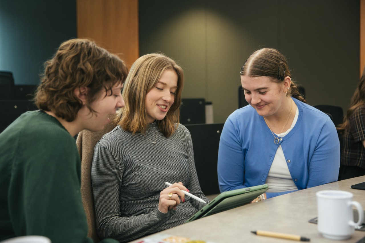 Three students sit together in a lecture hall. The student in the middle points at a tablet with a stylus.