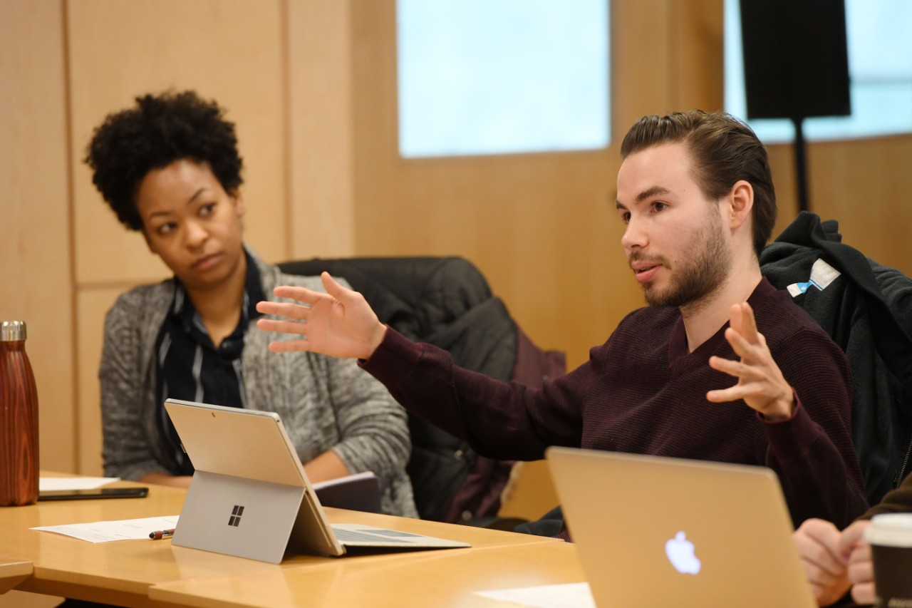 One person with long textured hair sits next to a second person sitting at table with a laptop with short brown hair and a short beard.
