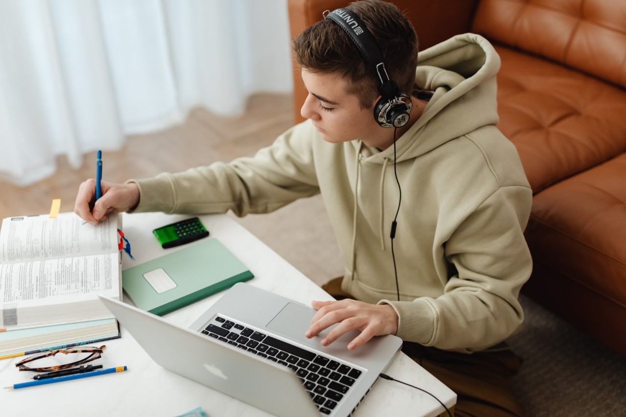 Student studying at latop wearing headphones and consulting text book.