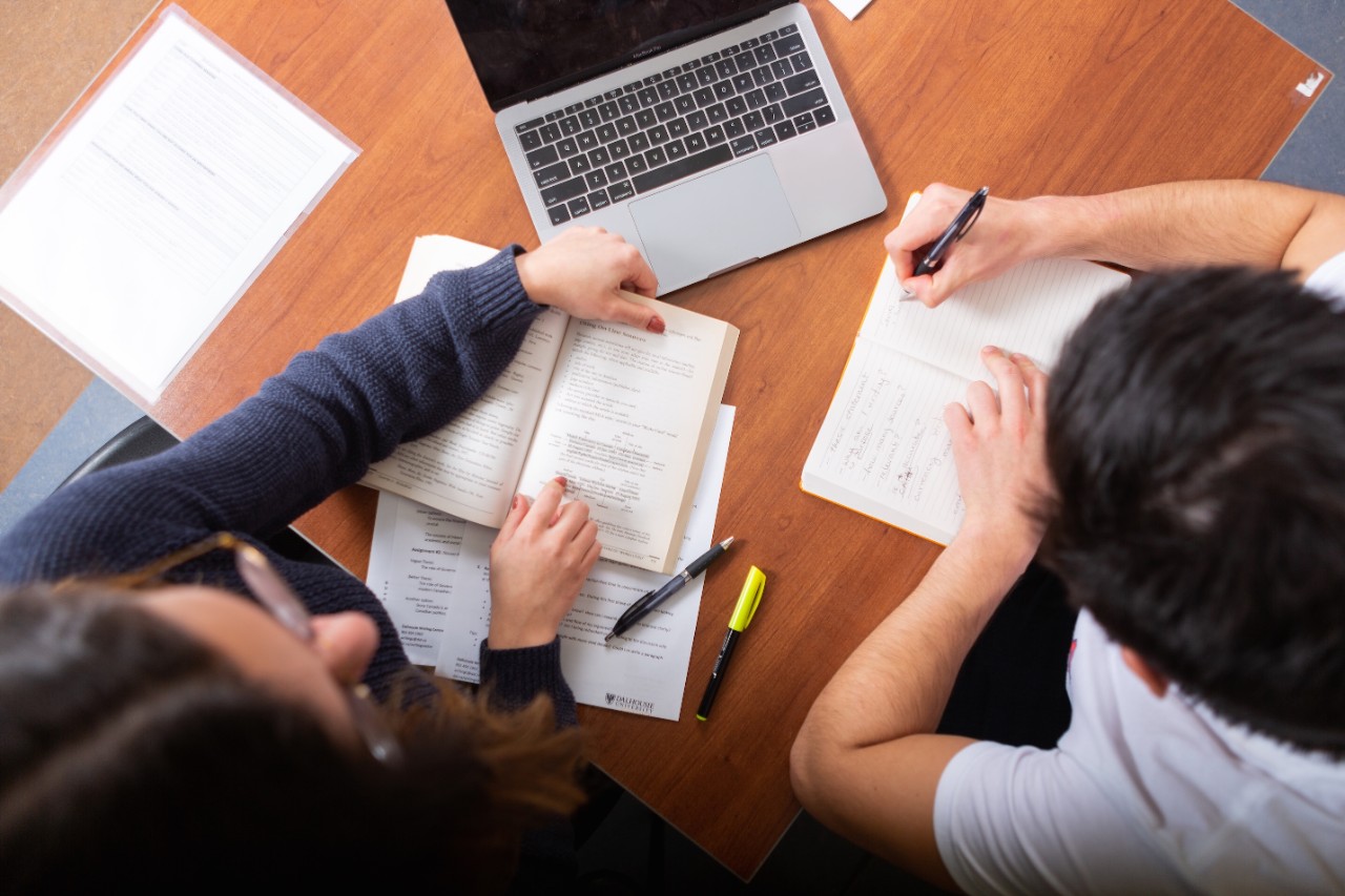 An overhead photo of an advisor and student working at a table with pens and notebooks. A laptop computer sits in front of them.