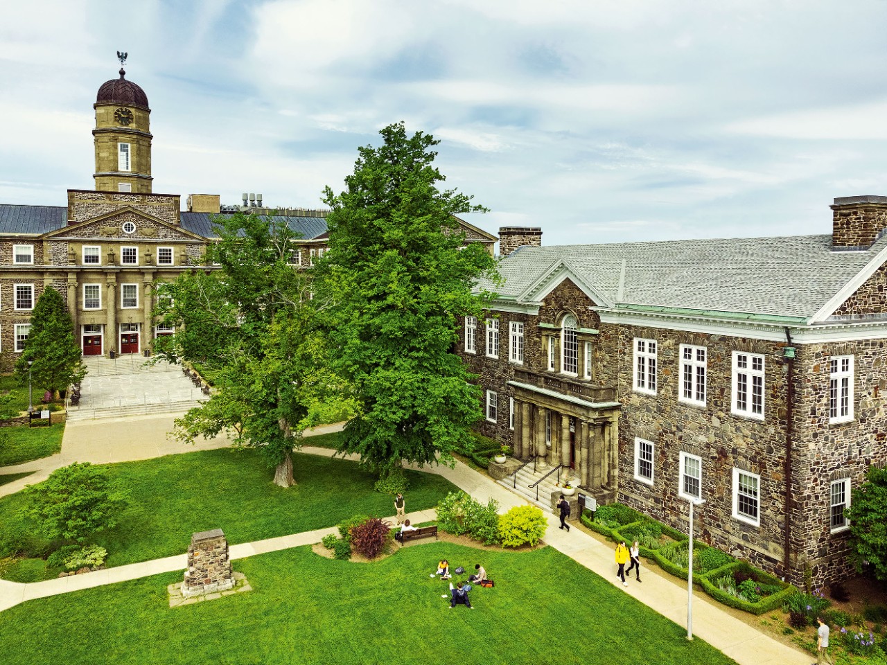 Students sitting and walking across green quad, with stone academic buildings in the background.
