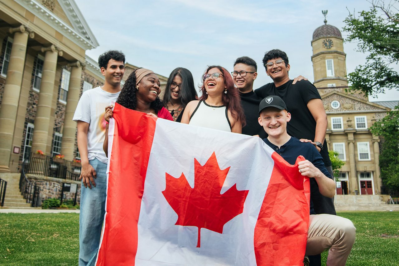 Seven students laugh while holding a Canada flag, posing for a photo in front of Dalhousie's Henry Hicks building.