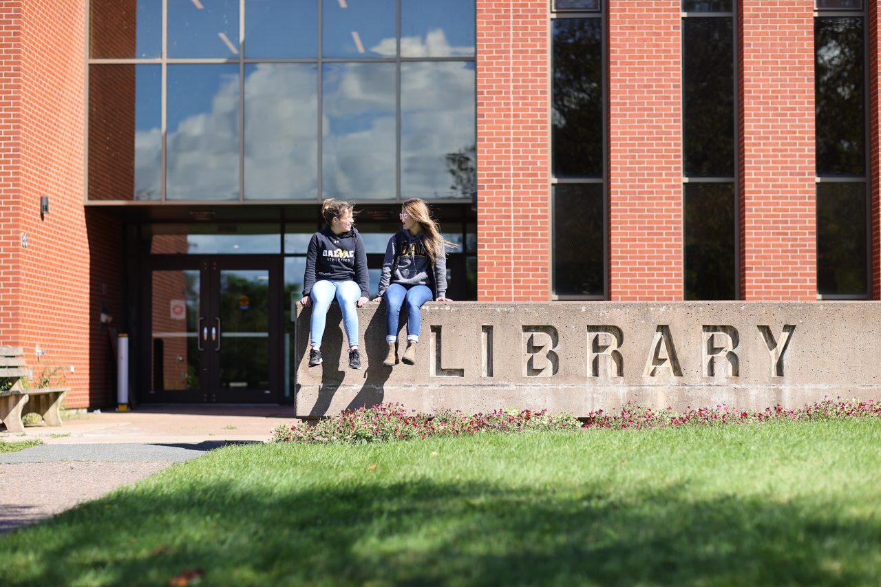 Two students sit on a large stone sign that says LIBRARY outside a red brick building. 