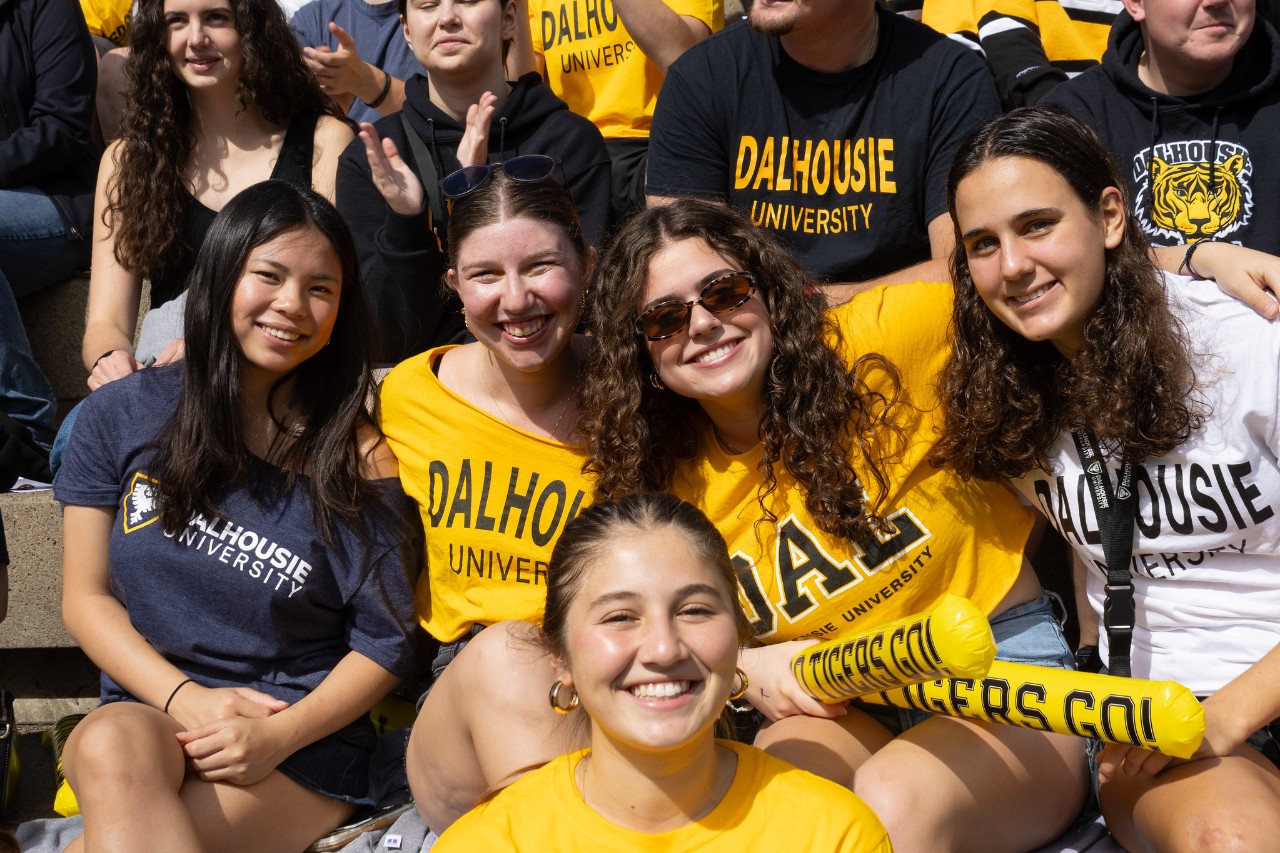 Five students wearing Dalhousie University t-shirts smile at the camera while sitting in a crowd of people.