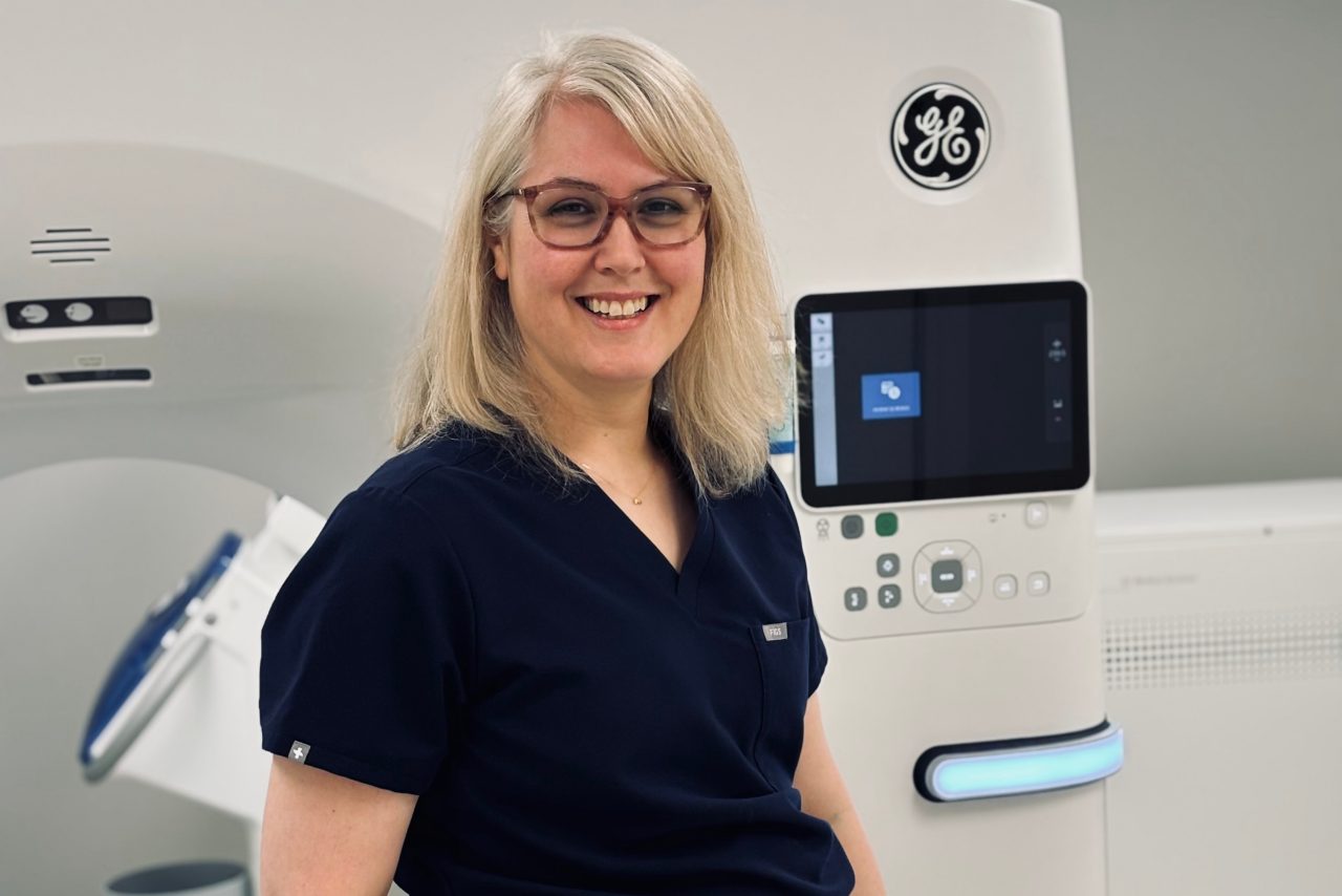 Leslie in scrubs, smiling in front of a CT machine.