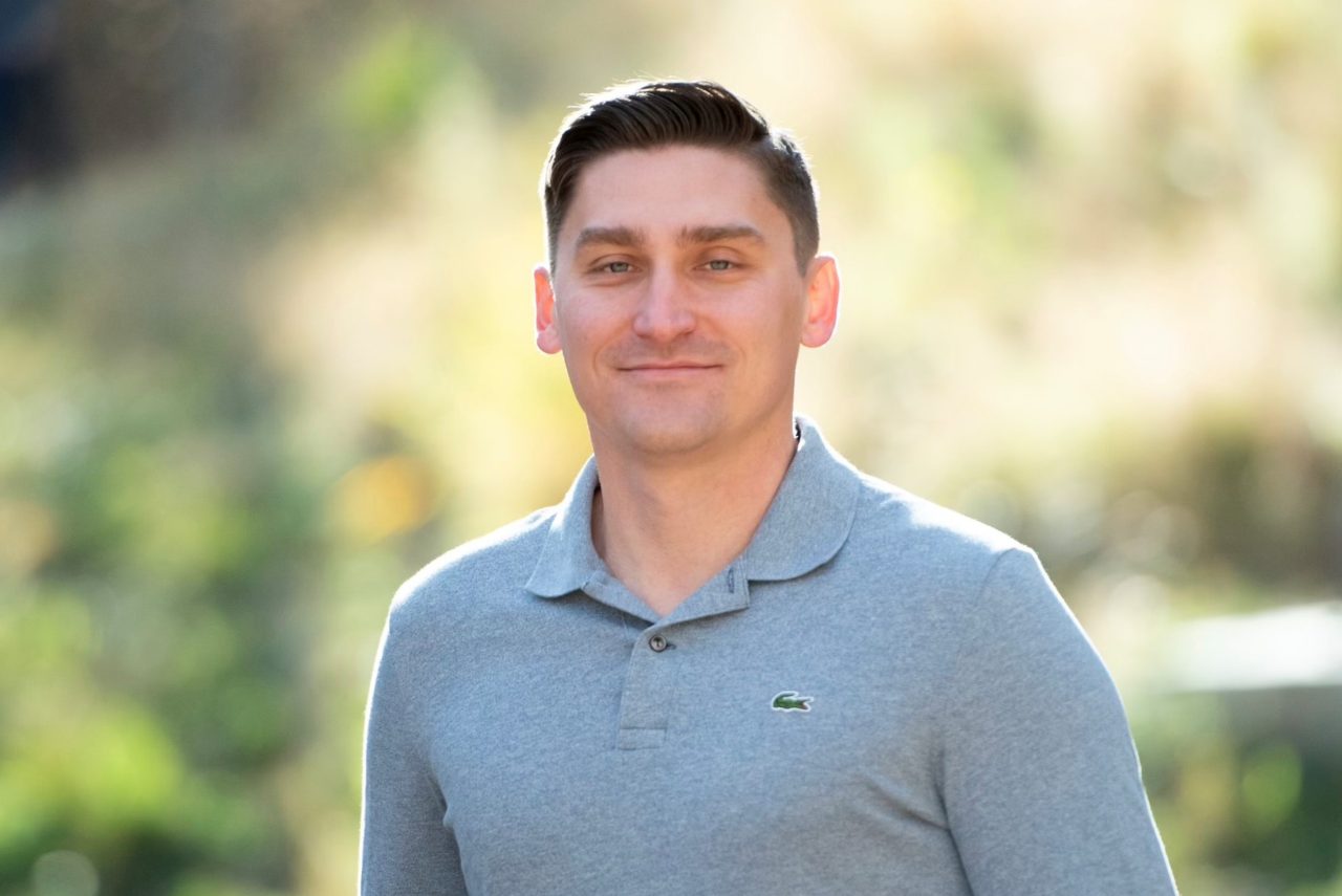 Stephen, wearing a grey collared shirt, stands in front of a backdrop of greenery. 