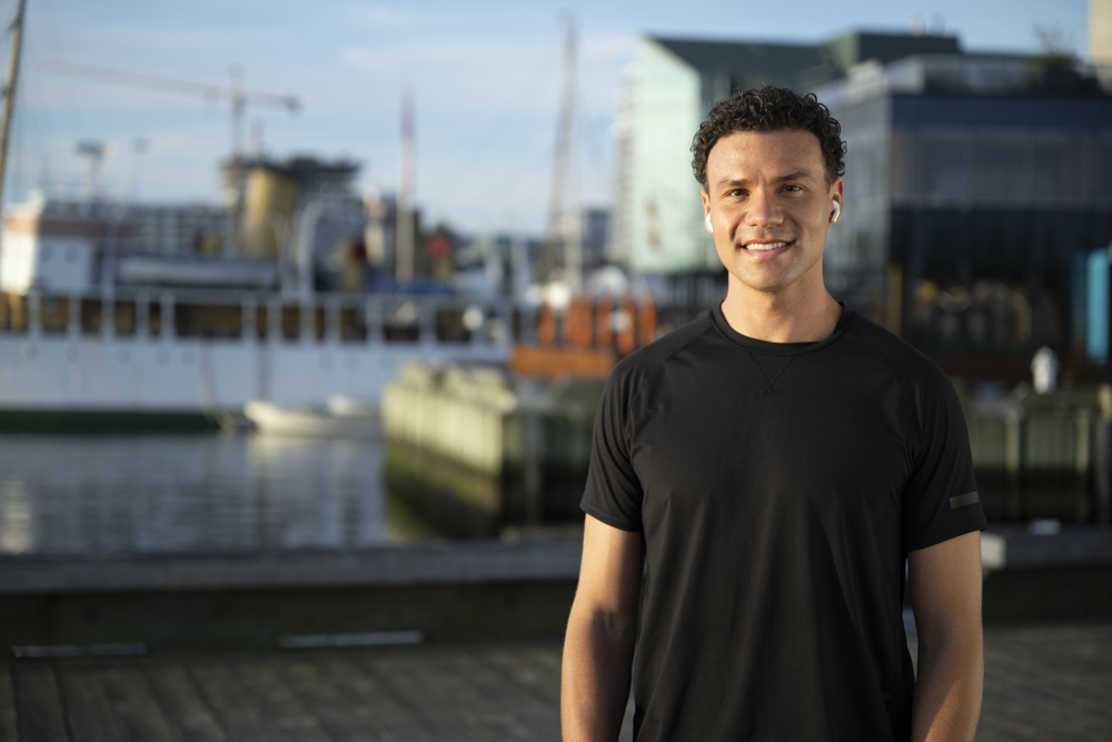 Daniel stands outside in an athletic black shirt, A harbour with boats is visible in the background.