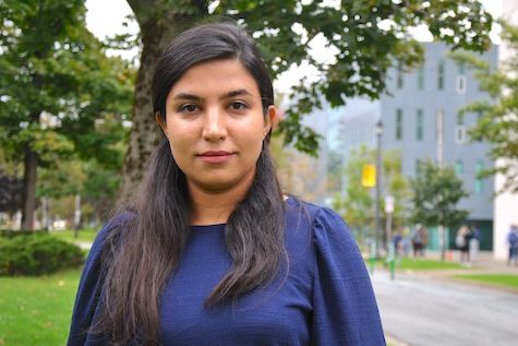 Reihane stands on a treed boulevard outside of the Rowe Building, wearing a blue shirt. 