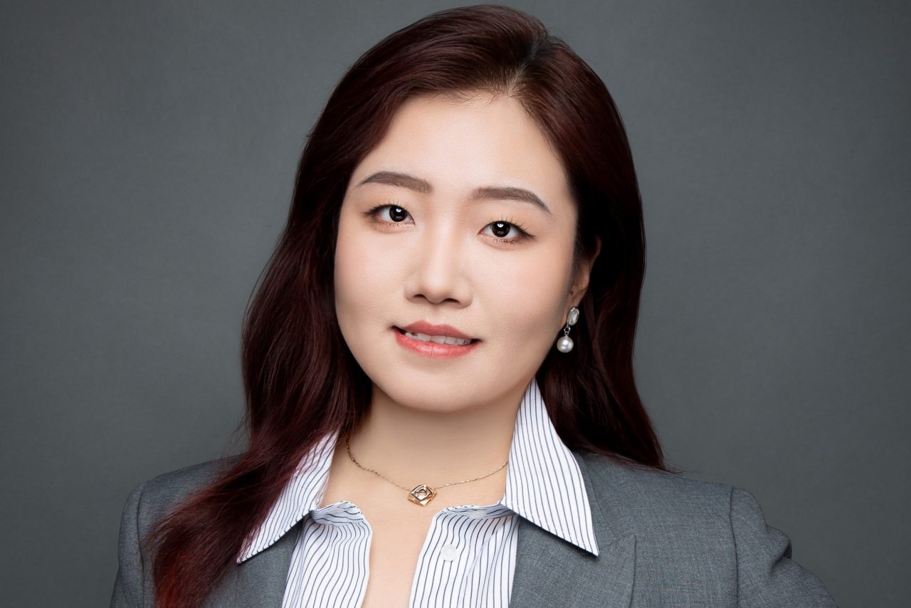 A portrait of Huiyan. She stands with one hand on her hip, wears a grey blazer and smiles. 