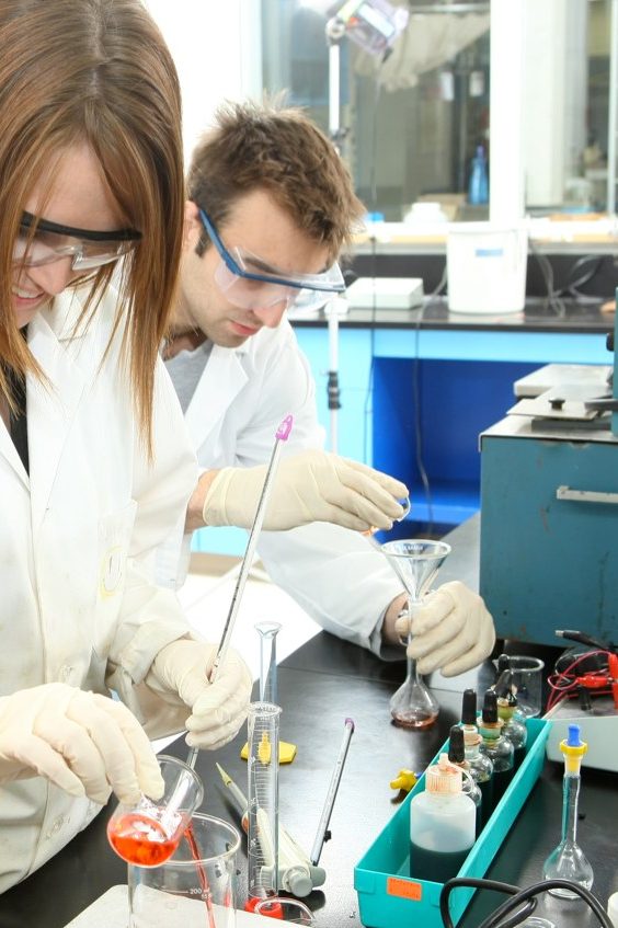 Three students in lab coats work at lab bench with various chemical equipment.  
