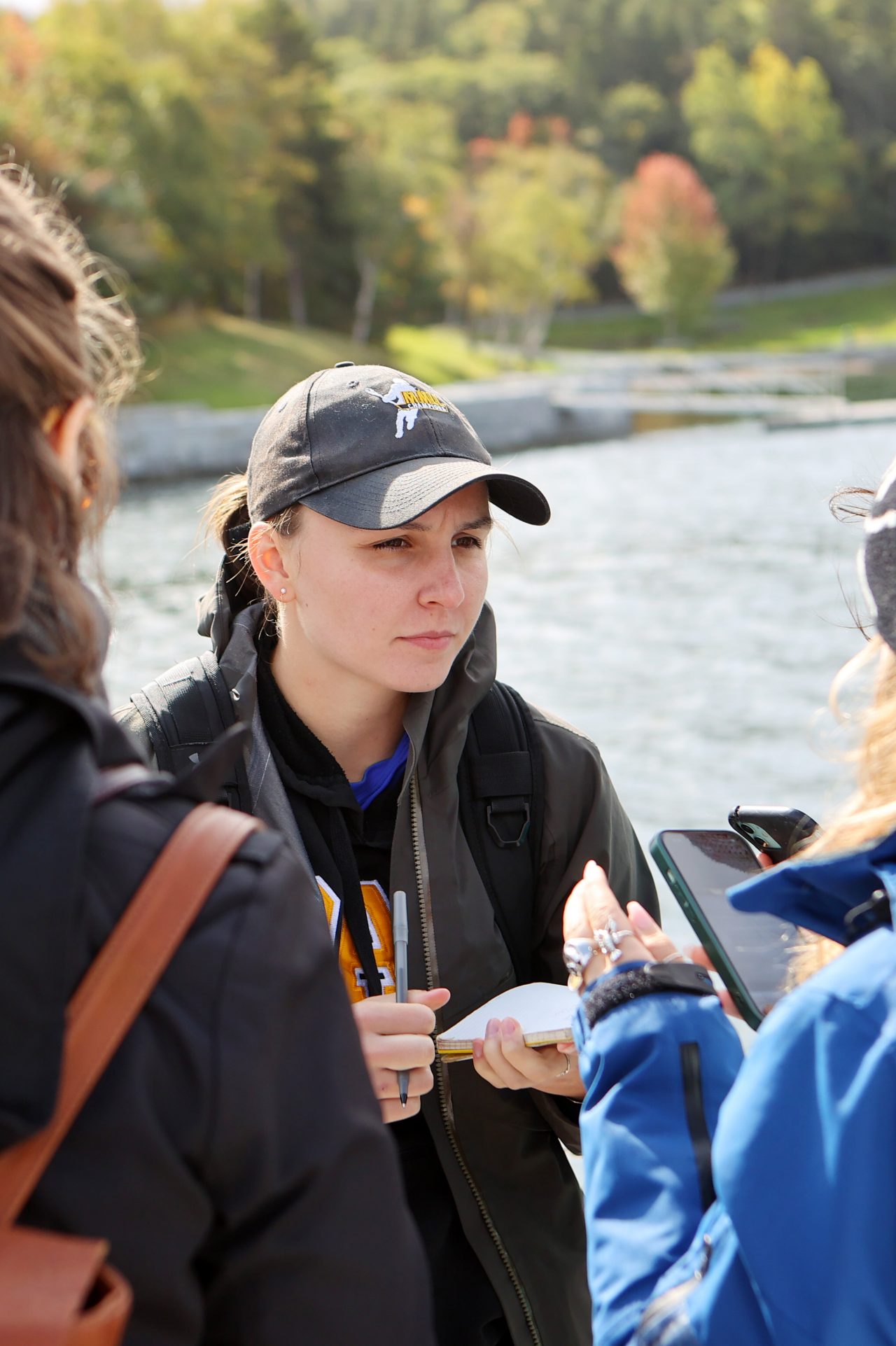A group of students dressed in hats and coats converse by the water's edge on a sunny fall day.