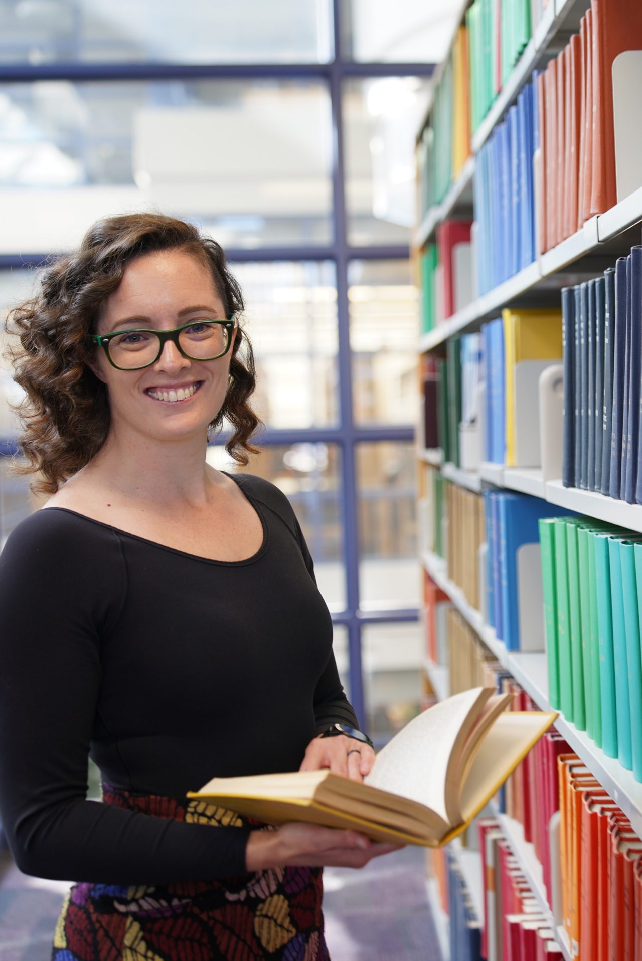 A person wearing a black shirt stands next to shelves filled with books, holding a book and smiling.