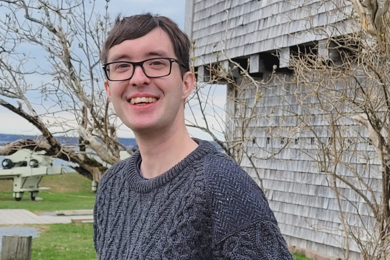 Keith stands outdoors, smiling in a grey cable-knit sweater. A grey building, trees and harbour are visible behind.