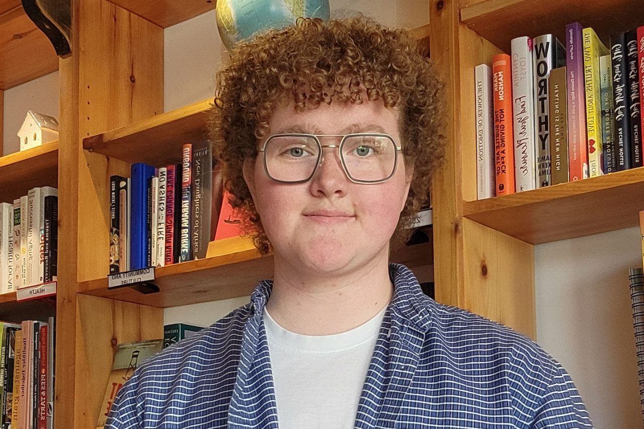 Rowan Moore stands in front of a bookcase, wearing a blue shirt.  