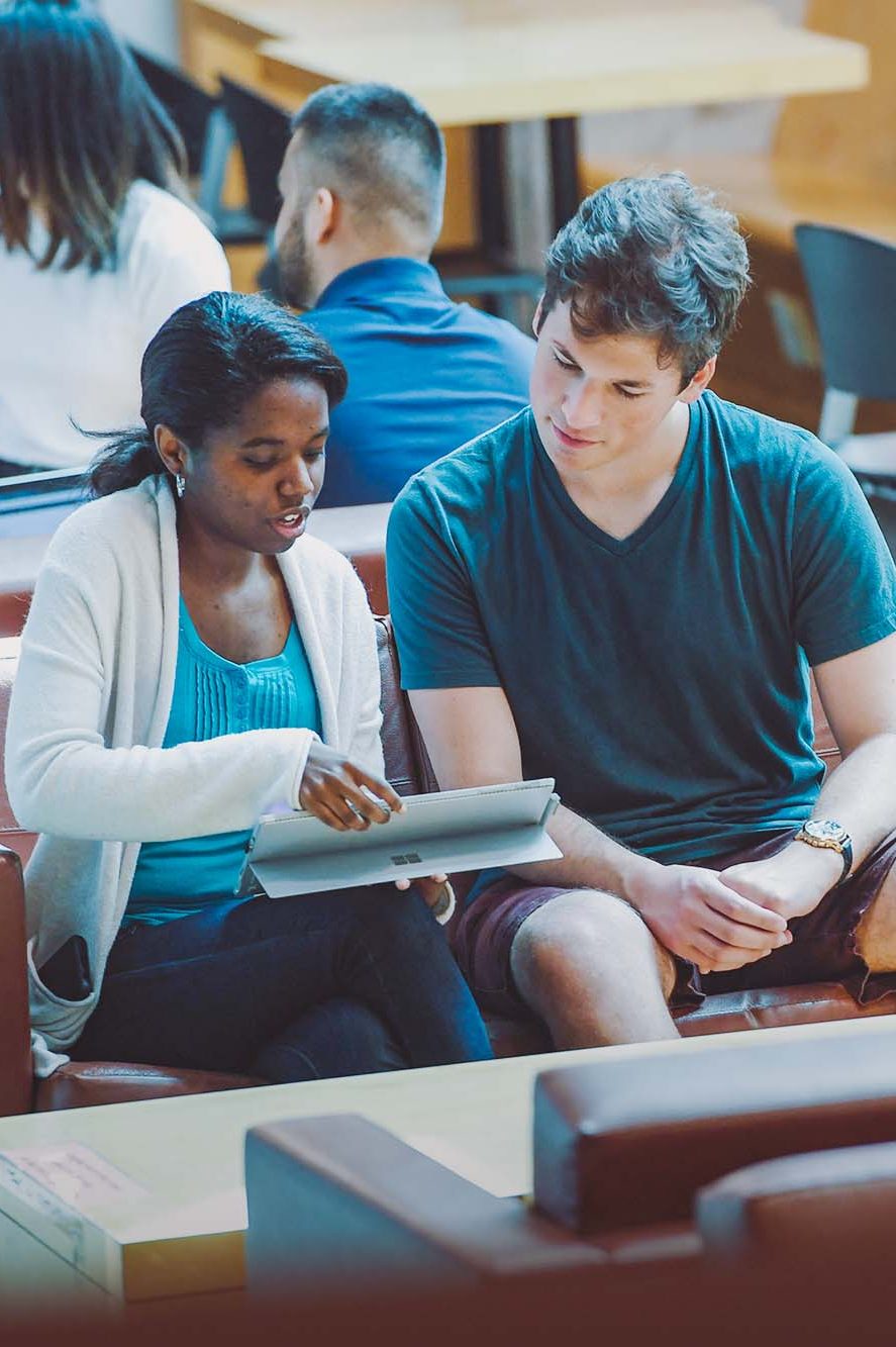 Students sit in a light-filled atrium, reviewing something on a tablet. 