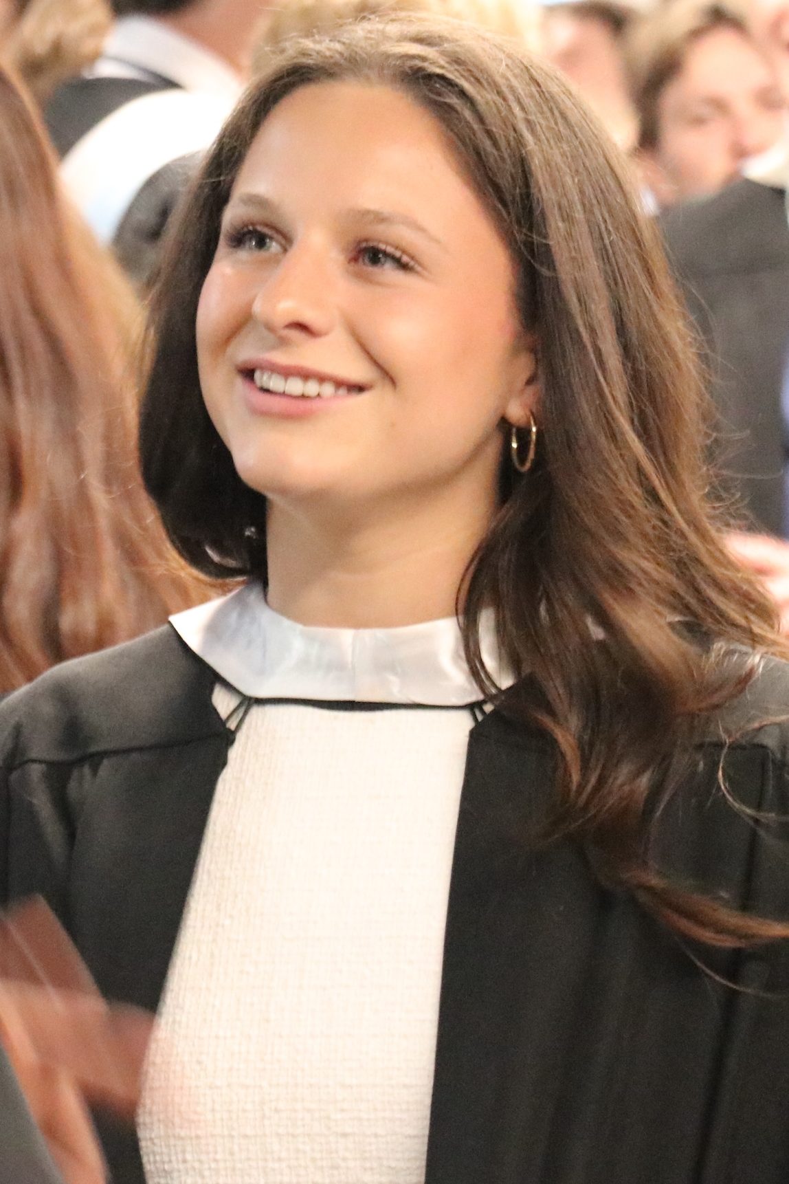 A student smiles at another student (facing away from camera). They are dressed in convcoation wear, with one wearing a kente sash in red, yellow, green and black.