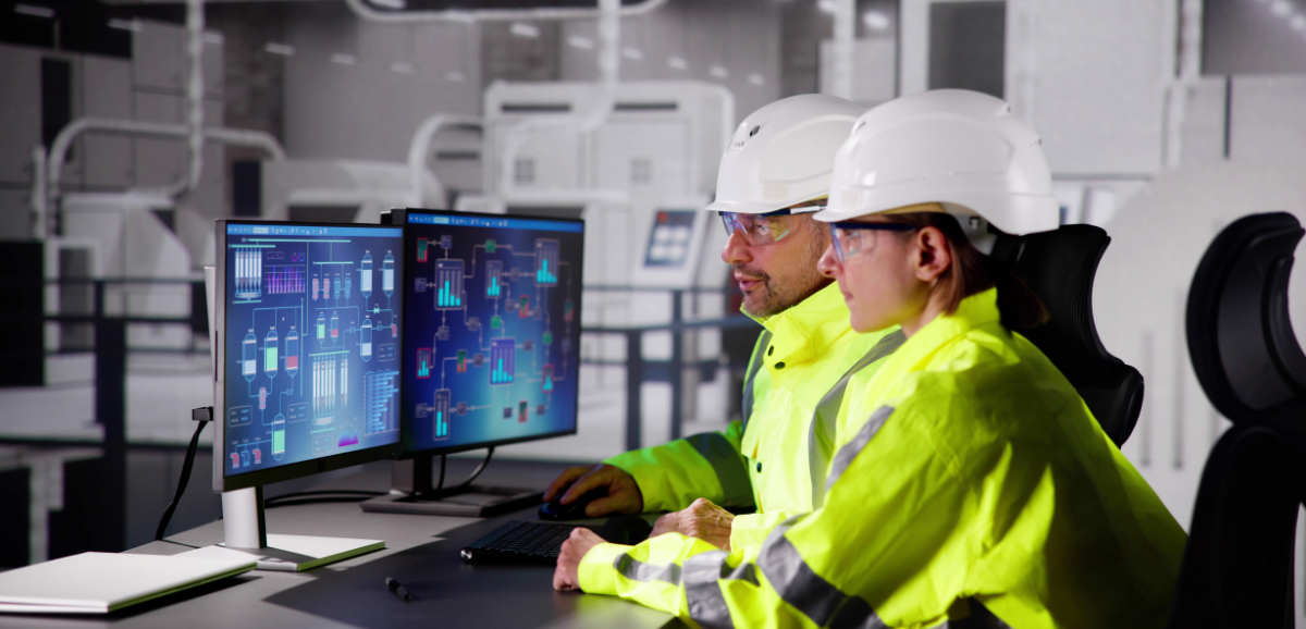 Two engineers wearing safety jackets and white hard hats sit at a desk looking at two computer monitors.