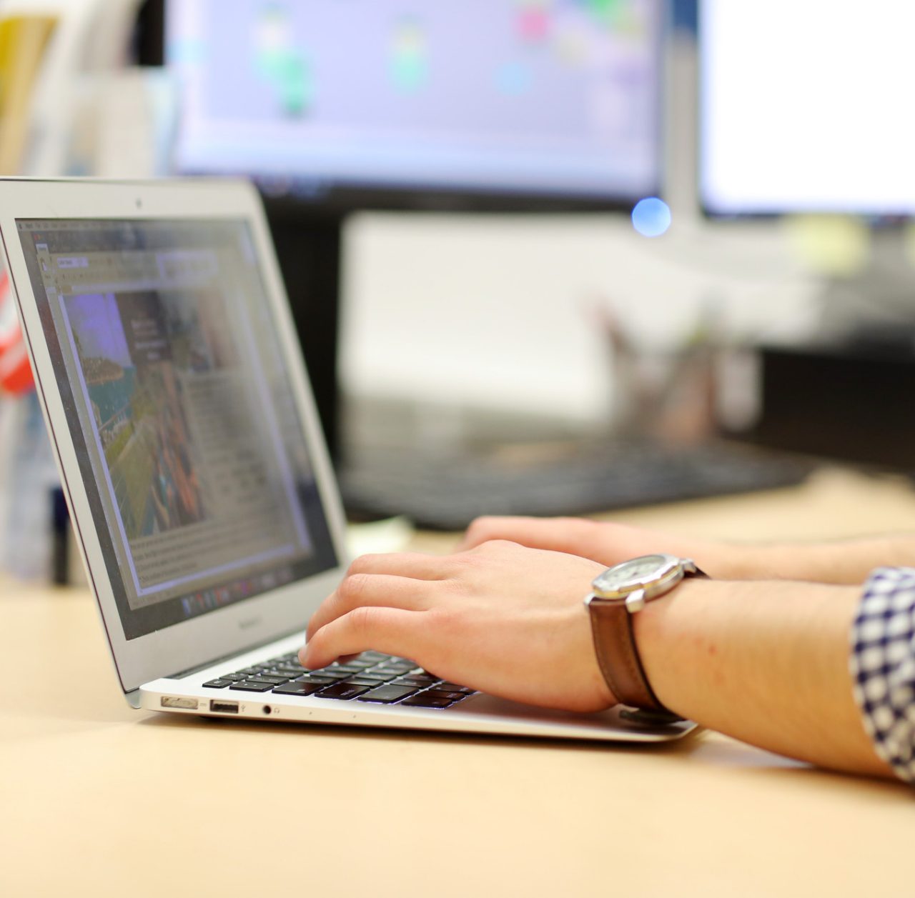 Close up of a person wearing a checked shirt and a brown watch typing on a laptop.
