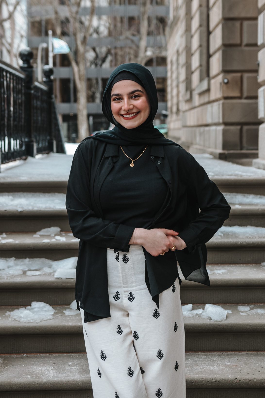 Mariam smiles, standing outdoors in front of some icy steps. She has a medium-light skin tone and is wearing a black hijab, black shirt, and black jacket over stylish white pants. 