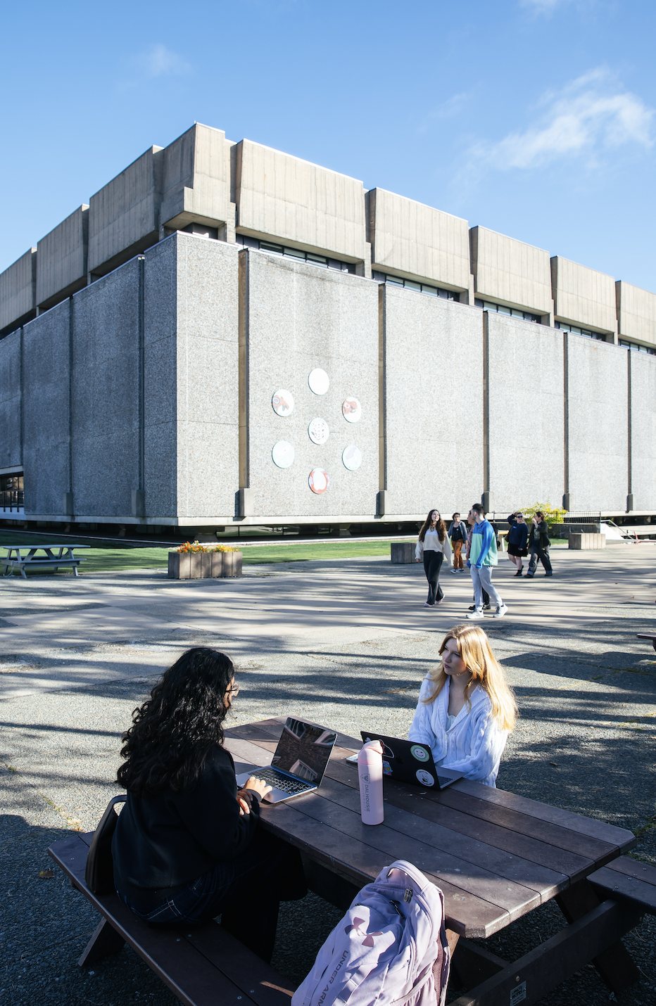 Two students sit doing work at a picnic table outside with library building in the background