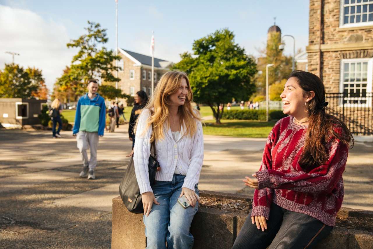 Two students sitting on a concrete bench chatting, with the Studley Quad in the background.