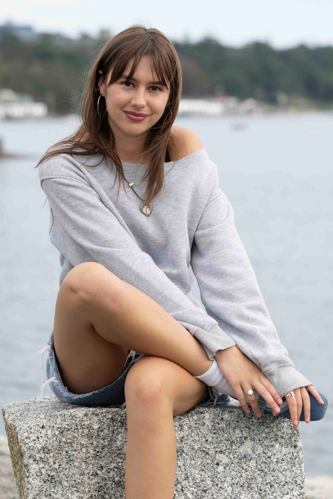 Abby sits on a rock facing the camera, smiling. In the background is water and the coastline. 