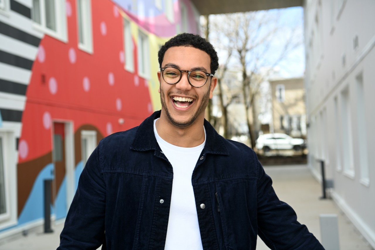 Mohammed, wearing a blue corduroy jacket, grins in front of a brightly-coloured mural. 