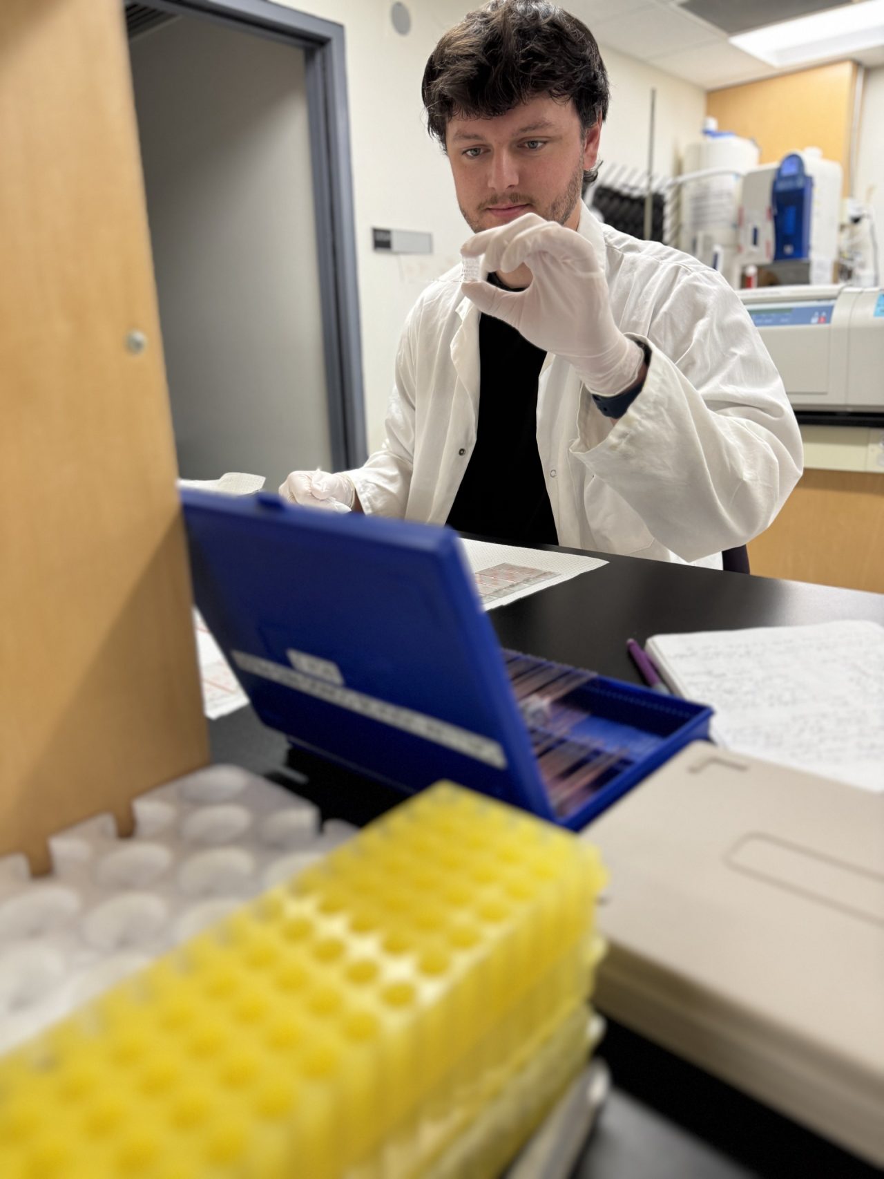 Male neuroscience student in lab coat inspects a small vial while wearing white gloves 