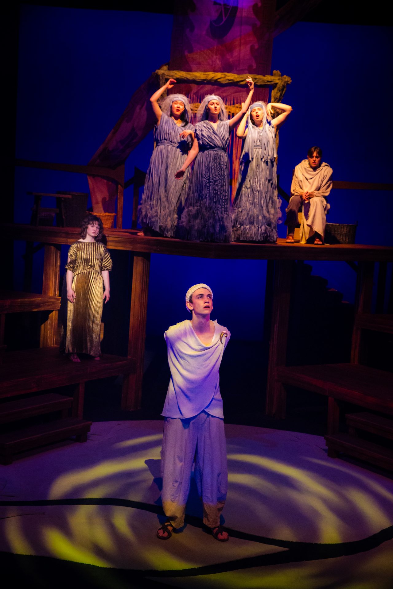 An actor stands centre stage with five actors on raised platforms behind them. They are costumed as Greeks and the lighting is blue and moody.