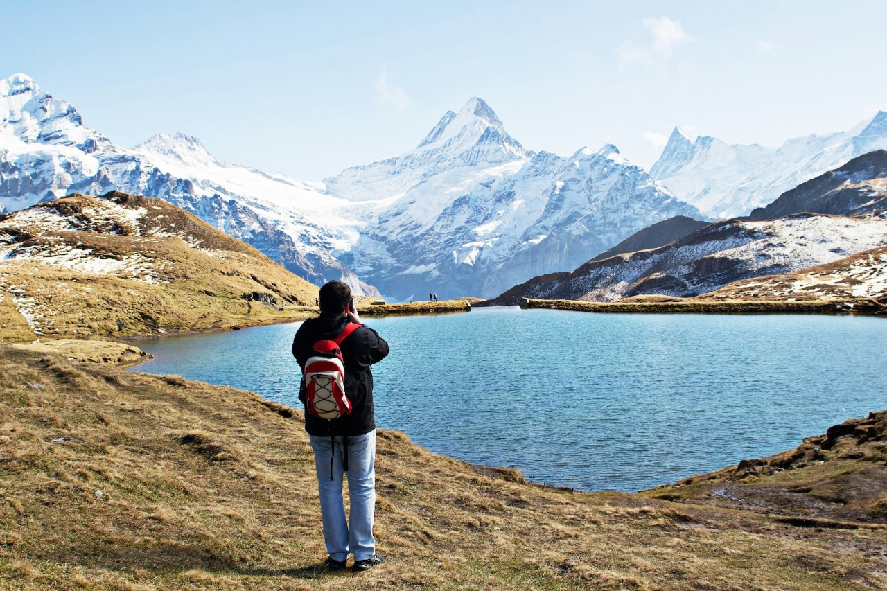 A student wearing a dark jacket and red and white backpack stands in front of a small lake with snow-covered mountains in the distance.