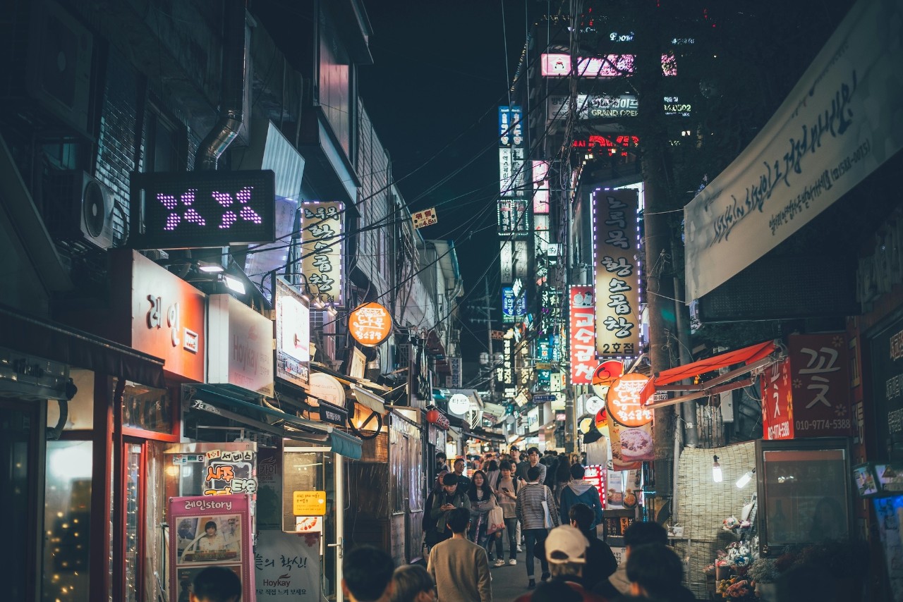 A busy narrow street at night is filled with bright signs with Korean writing.