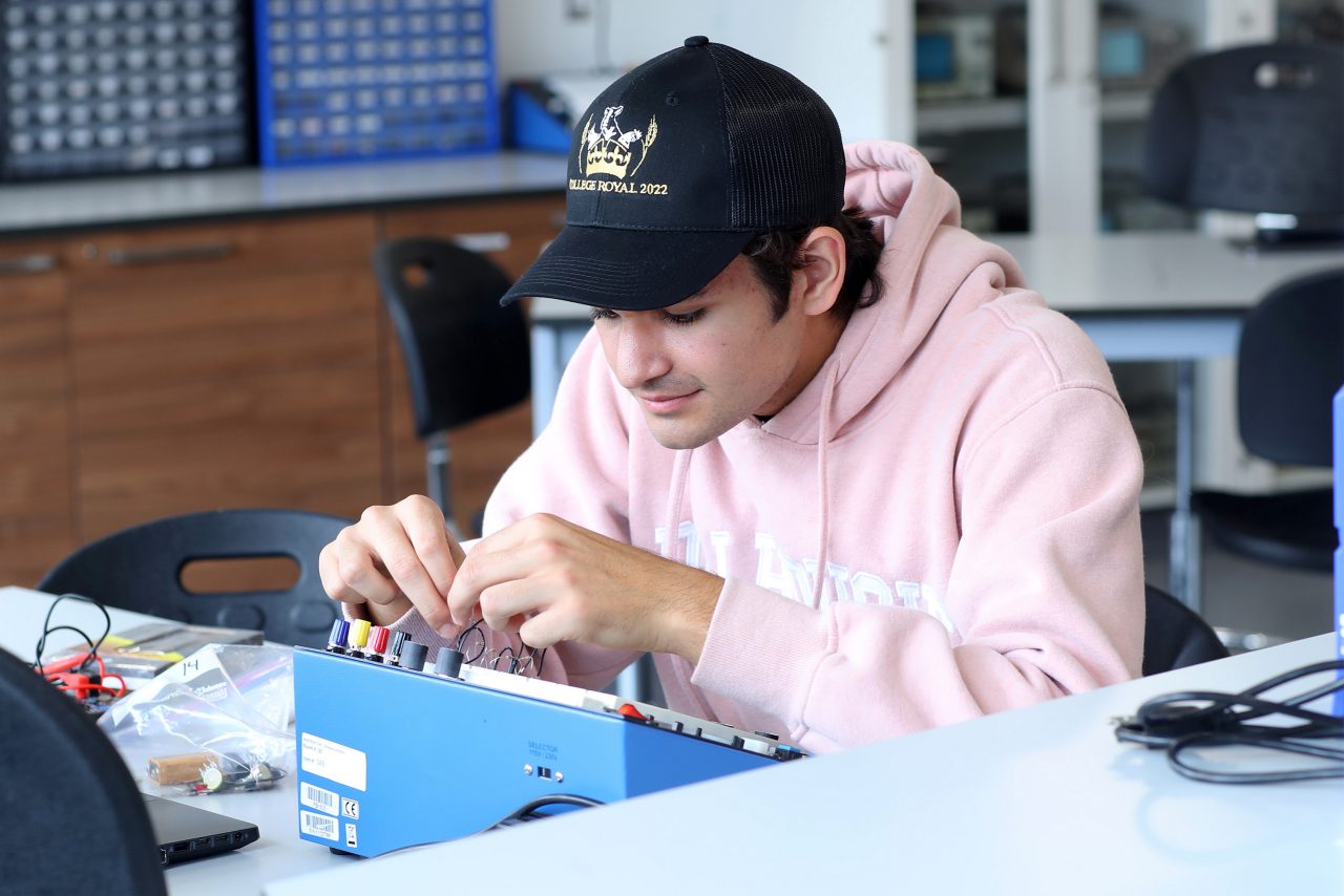 A student wearing a pink hoodie and black baseball cap adjust dials on a blue box.