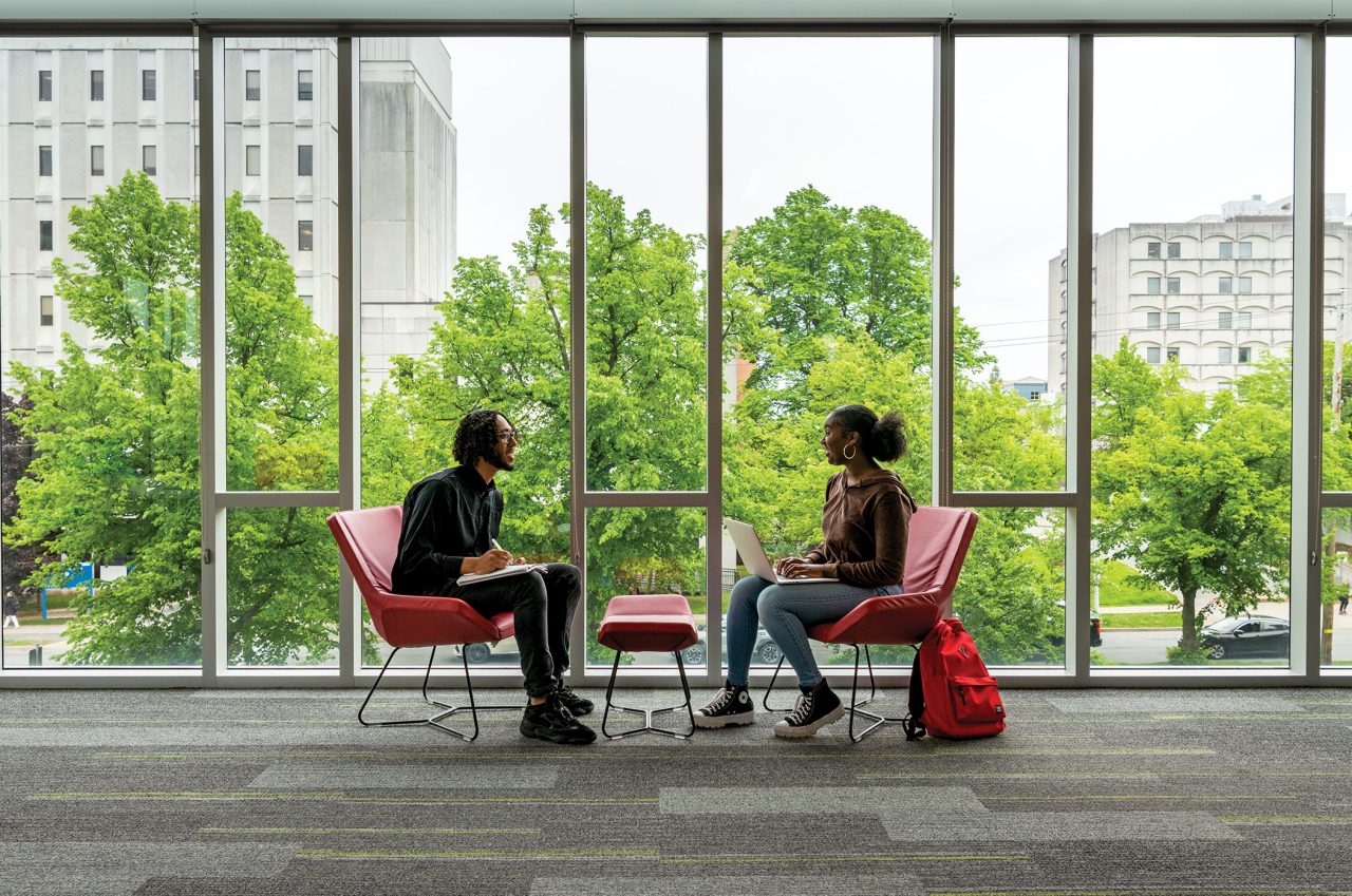 Two students sit by a wall of windows